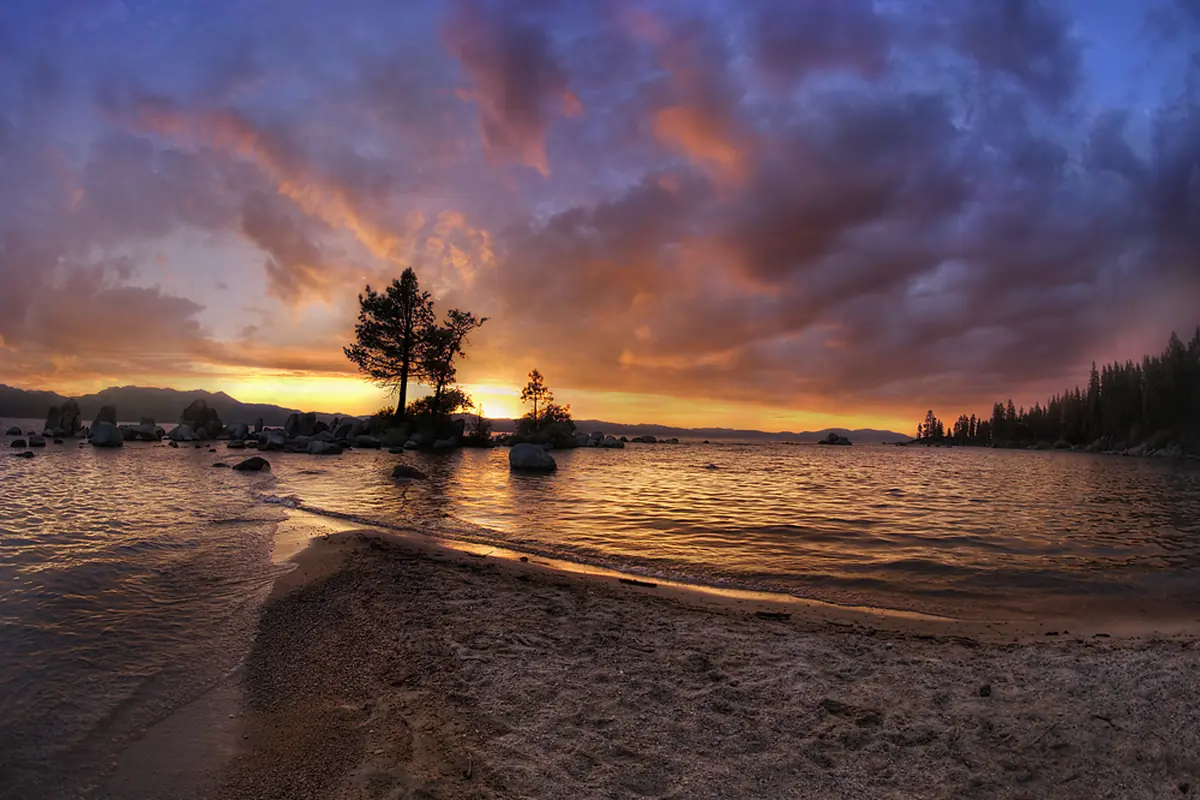 An orange and violet sunset over Lake Tahoe seen from a sandy shore at Zephyr Cove, with a small pine on a rock silhouetted against the glow